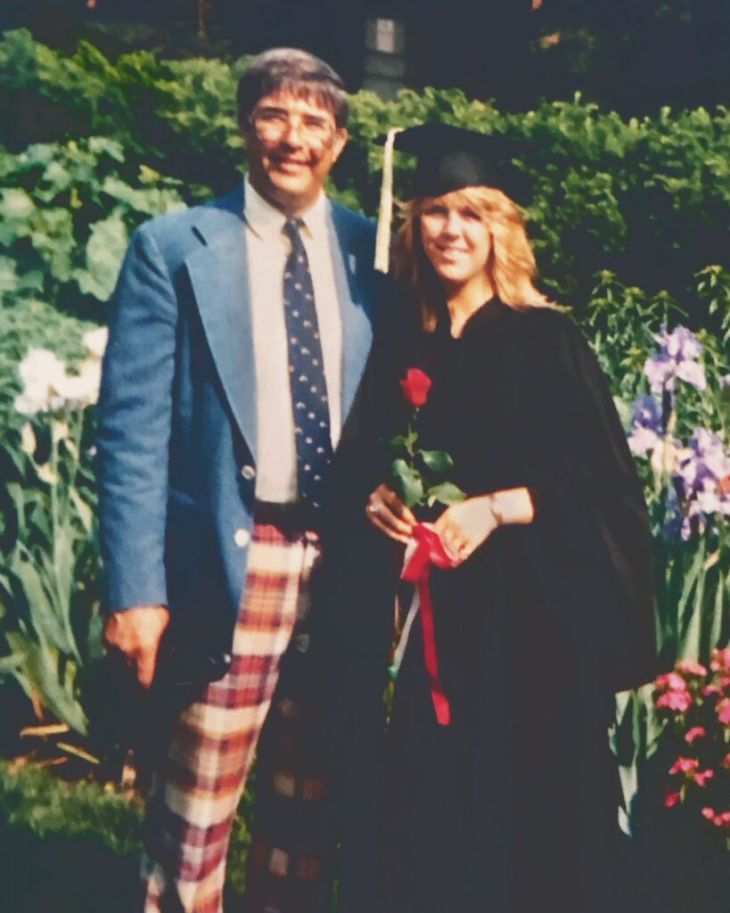 A woman in a black graduation cap and gown poses with an older man wearing a blue blazer and red plaid pants.