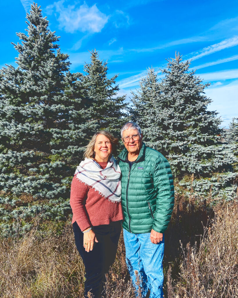 A man and a woman stand in front of a group of tall evergreen trees.