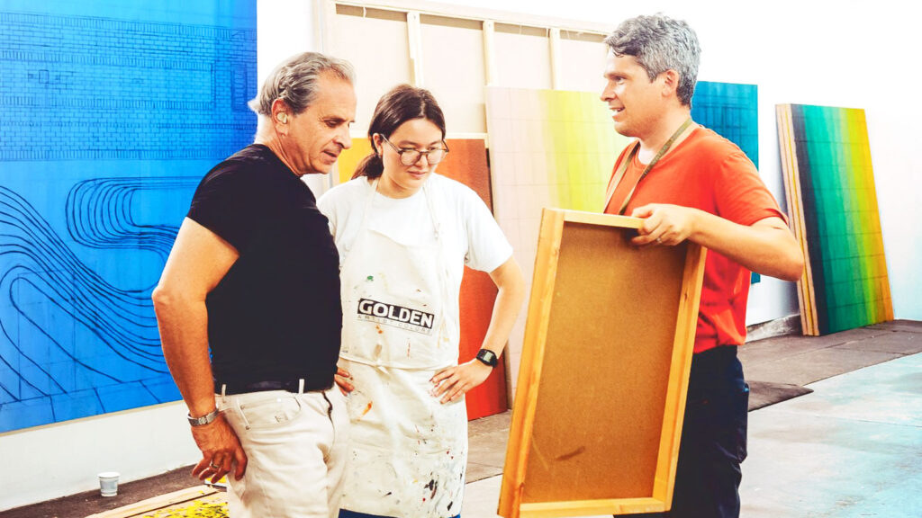 Two men and a woman stand around a painting being held by one of the men in an art studio