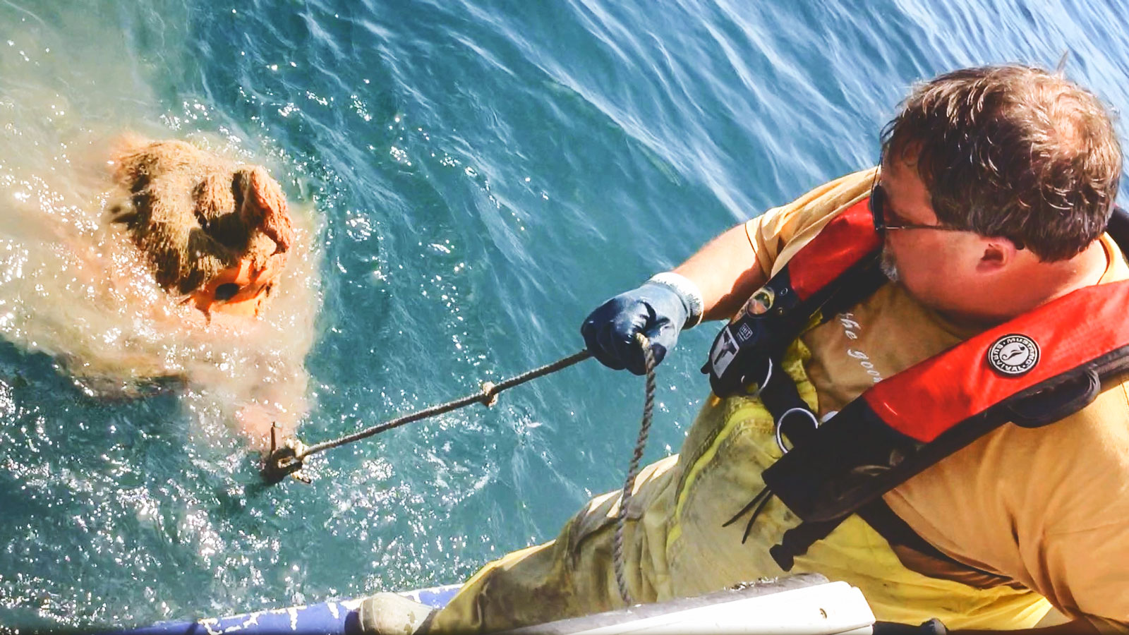 A man pulls a large orange audio recording unit out of the water with a rope while standing at the edge of a boat.