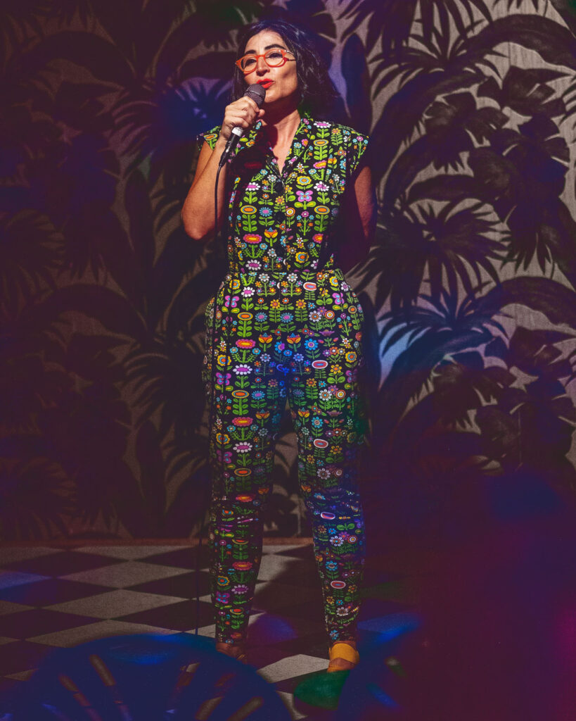 Comedian Negin Farsad performs standup comedy in Brooklyn wearing a patterned green jumpsuit.