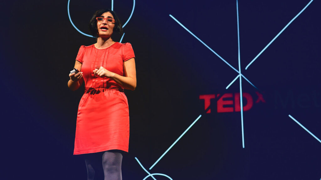 Comedian Negin Farsad gives a TED talk in front of a dark background with the word TED illuminated in red, while wearing a bright red shirt and glasses.