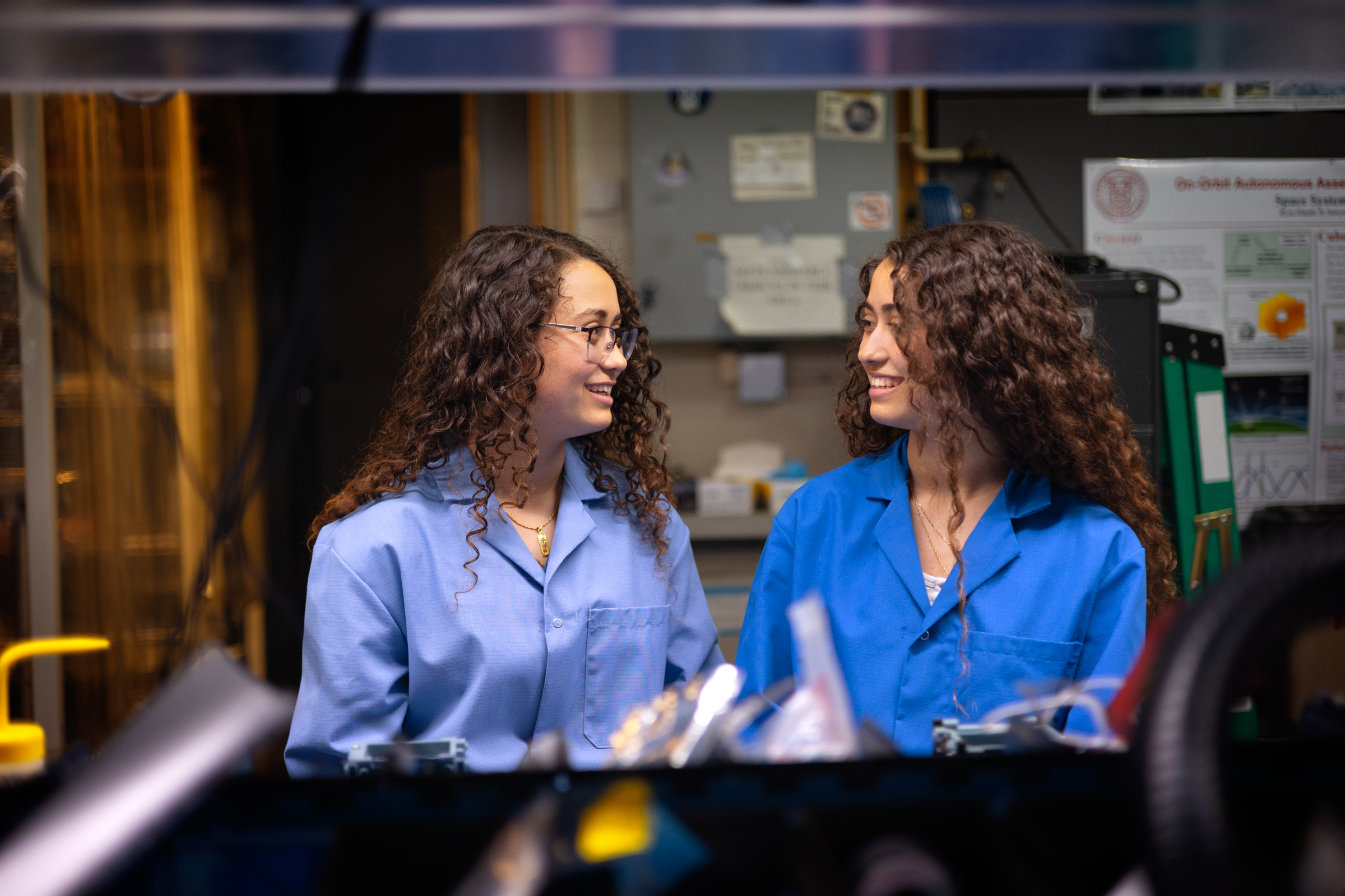 Twin sisters and mechanical engineering majors Ashley (L) and Verena (R) Padres, both class of 2026, chat at Verena's workbench in the Space Systems Design Studio Lab, Rhodes Hall.