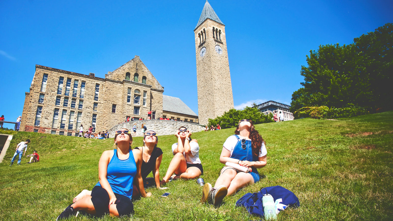 Four women wearing eclipse glasses sitting on the Slope with the tower in the background