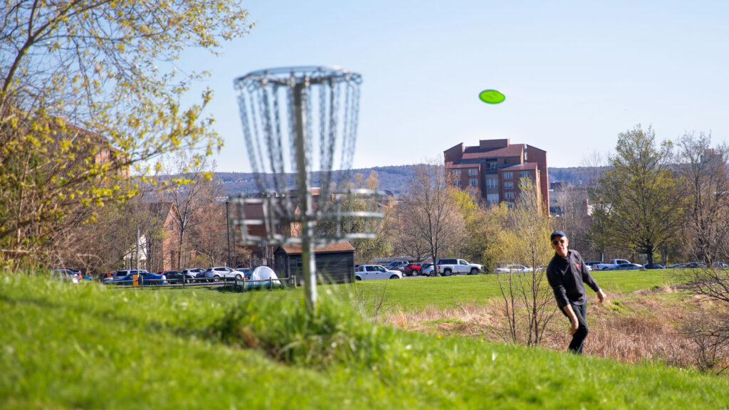 Bill Nye throws a Frisbee at a disc golf goal on the Cornell campus.