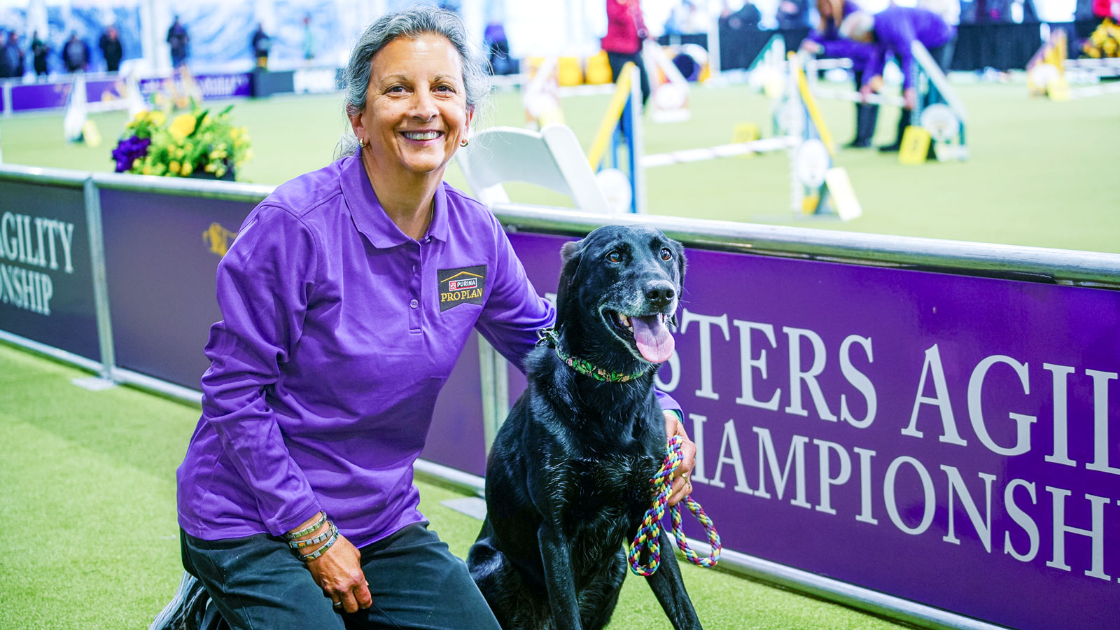 A woman in a purple shirt kneeling next to a black dog next to an agility course