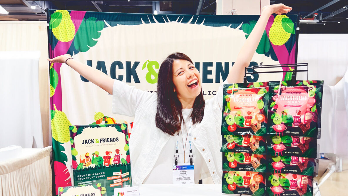Jessica Kwong smiles and throws her hands in the air in front of her vegan jerky products