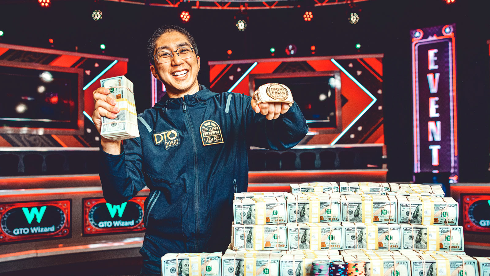 Jonathan Tamayo holding his bracelet trophy and a stack of cash, behind a large stack of cash