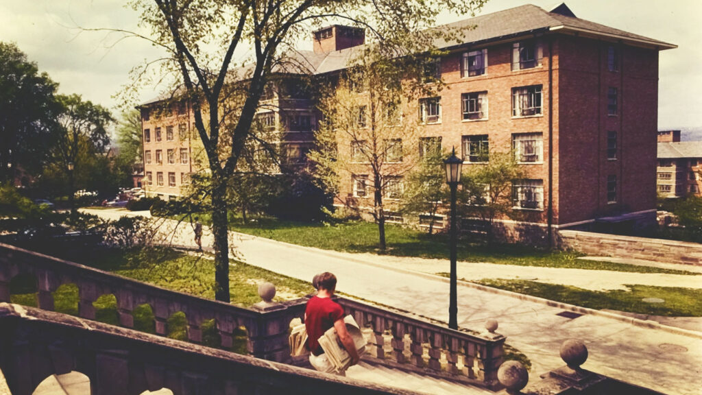 A student carrying newspapers descends stairs on West Campus with a University Hall visible across the street, 1967