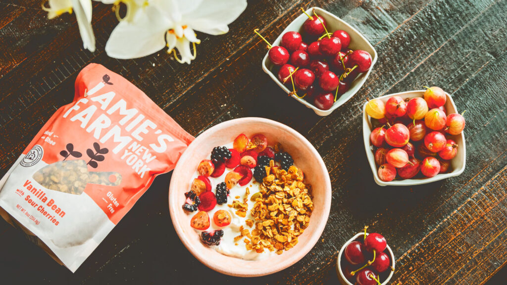 Jamie's Farm Vanilla Bean with Sour Cherries bag of granola sits on a wooden table with granola and cherries