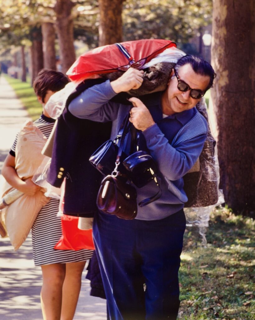 A dad carries clothing on hangers and several purses and handbags in a scene from Move-In on campus, 1970