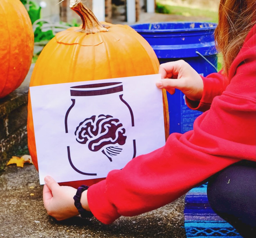 a stencil for carving a brain-in-a-jar design on a pumpkin is held up against a pumpkin