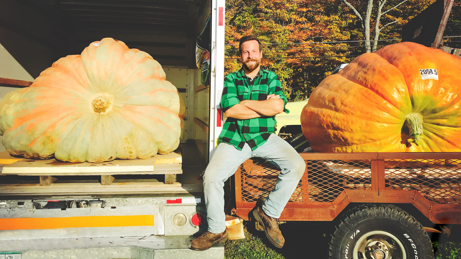 Alex Noel smiles while sitting on a wagon with two of his giant pumpkins to either side of him.