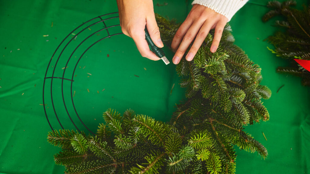 Hands wrapping a bundle of greenery to a metal wreath frame with wire.