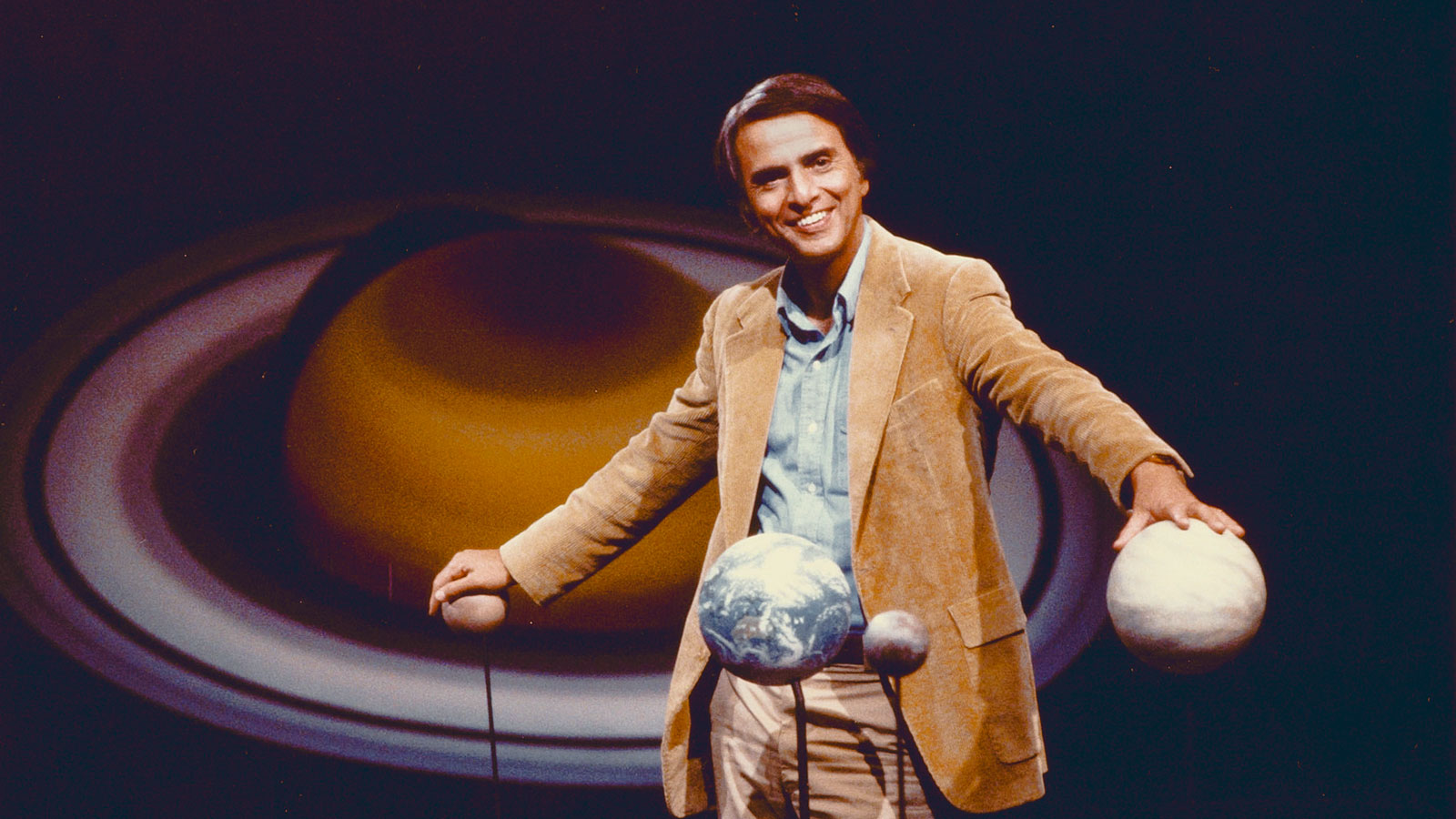 Carl Sagan pictured with models of the planets and a large image of Saturn behind him