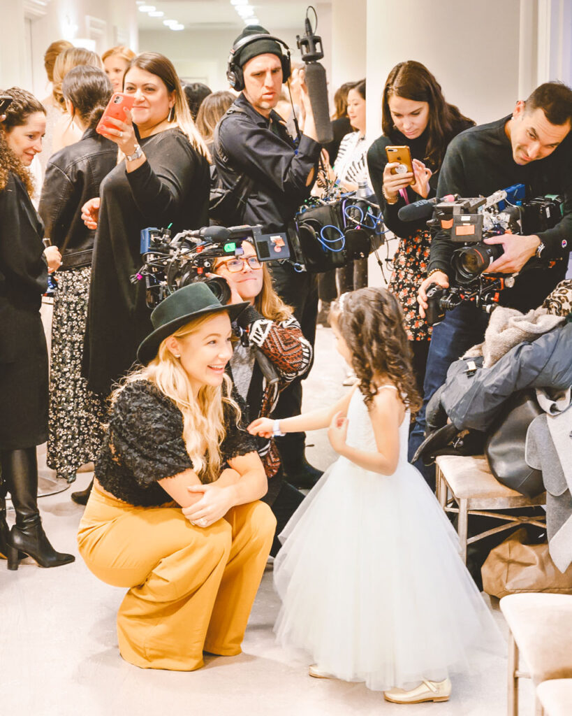 Hayley Paige kneels to meet a young girl wearing a white dress at eye level. There are photographers and videographers in the background documenting the moment.