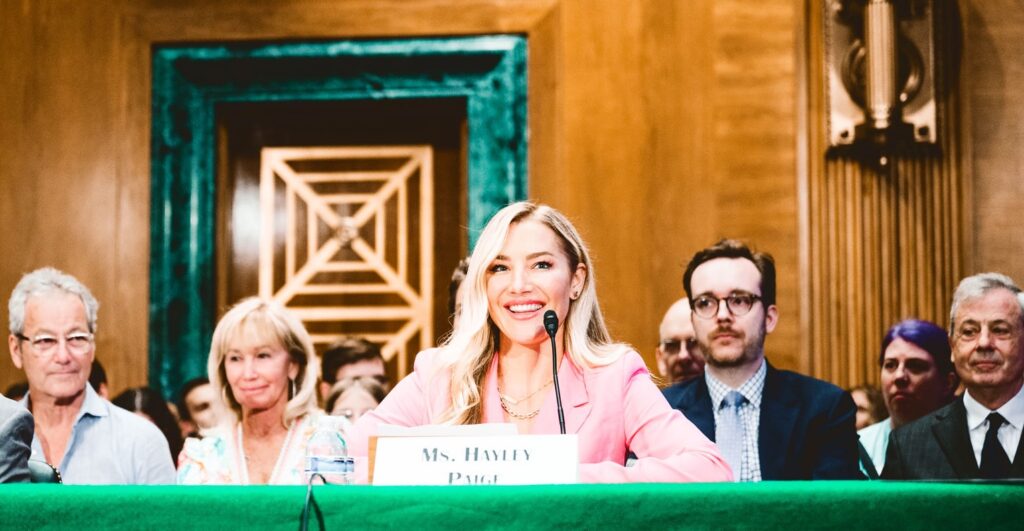 Hayley Paige sits with a microphone at a table while talking to Congress about her experience.