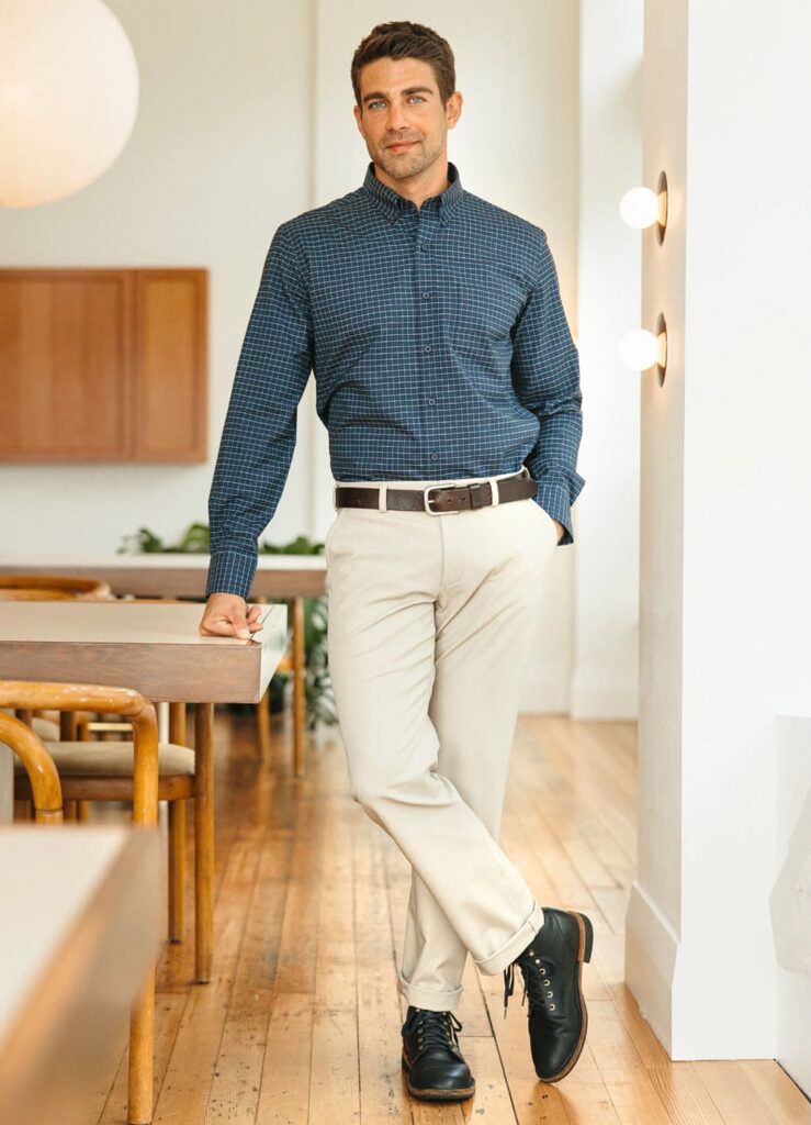 A male model standing wearing a blue checked dress shirt and light tan khaki pants