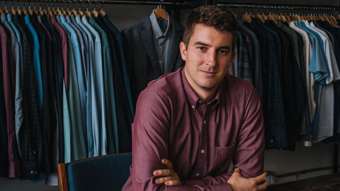 Mac Bishop wearing a maroon button down shirt in front of a rack full of dress shirts and other shirts