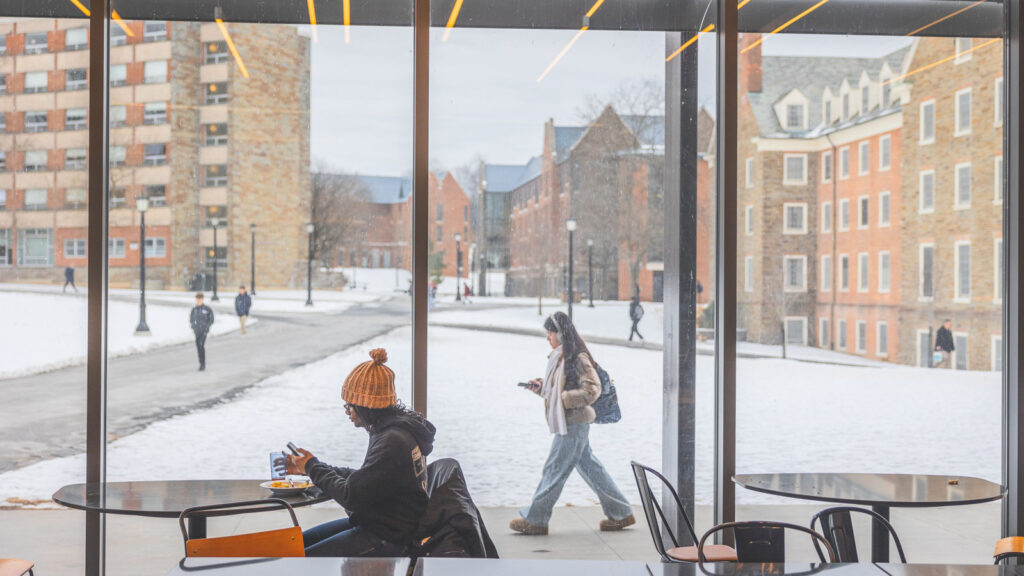 A student at a ground-floor table at a North Campus residence as students are seen through the windows walking along the paths outside in winter