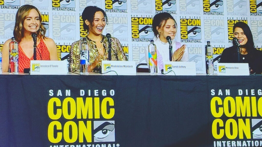 Jessica O'Toole sits at a table with a microphone and name card in front of her with three cast members from the Charmed series at Comic Con in San Diego.
