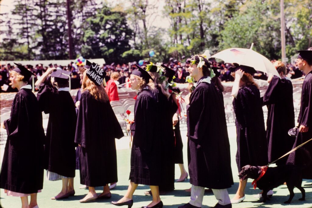 Grads in robes; one is wearing a cap that says "job wanted"