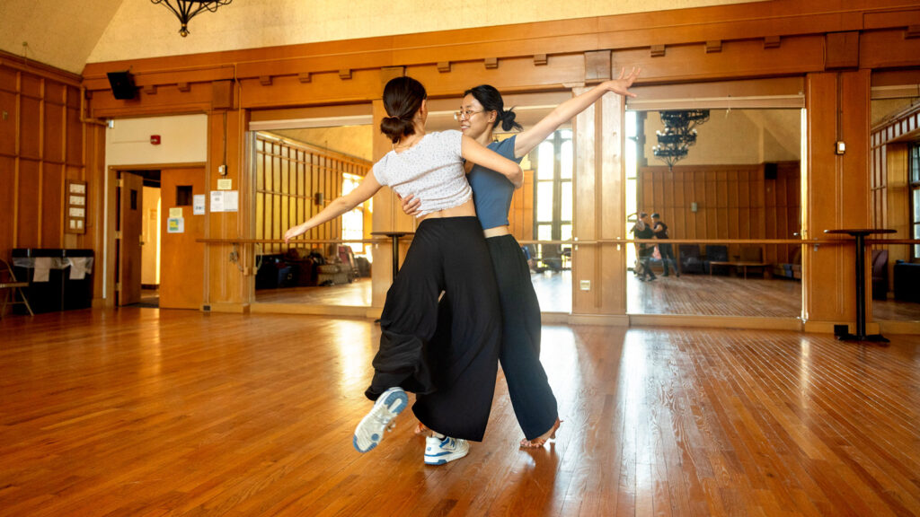Two girls rehearse a dance together in Willard Straight Hall with one arm around each other and one arm extended outward.