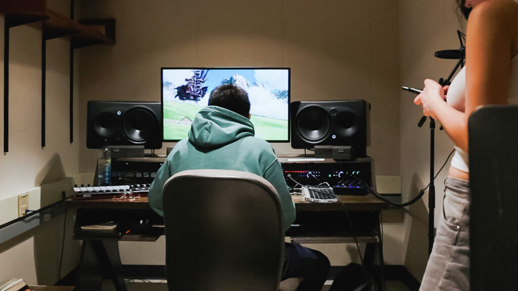 A student works at a desk with a computer monitor, music production equipment, and speakers in front of them.