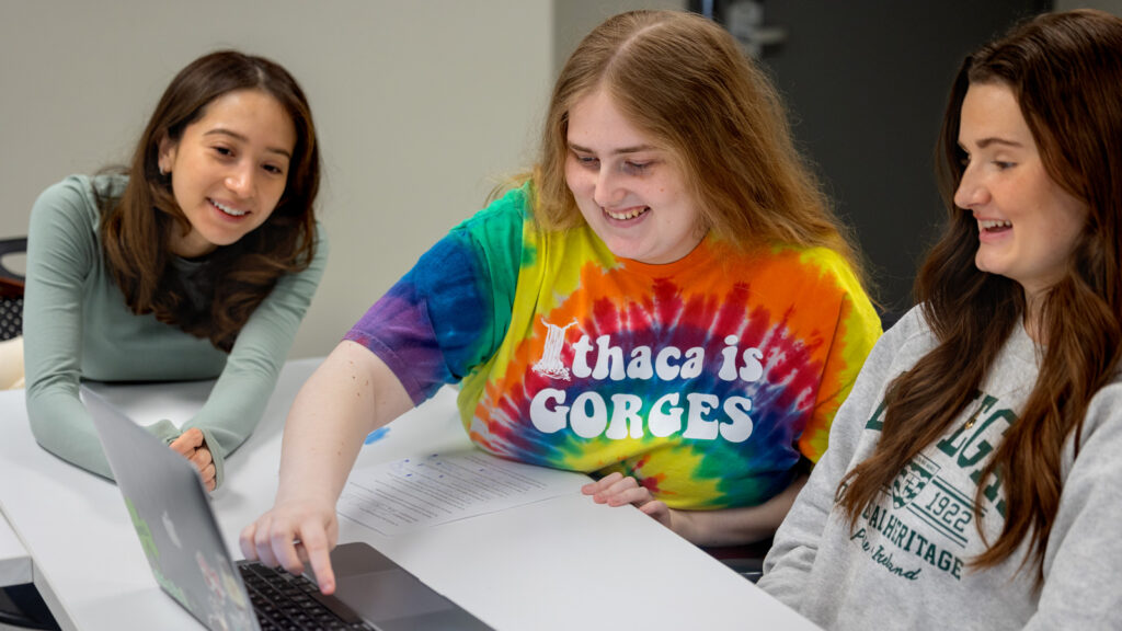Two Cornell students help a high school student with homework in front of a laptop.