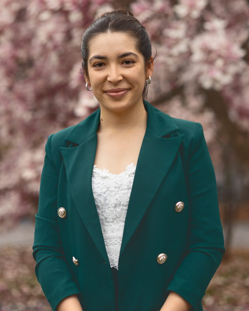 Diana Ayubi smiles in front of a light pink flowering tree in the Arts Quad while wearing a green blazer and white lace shirt.