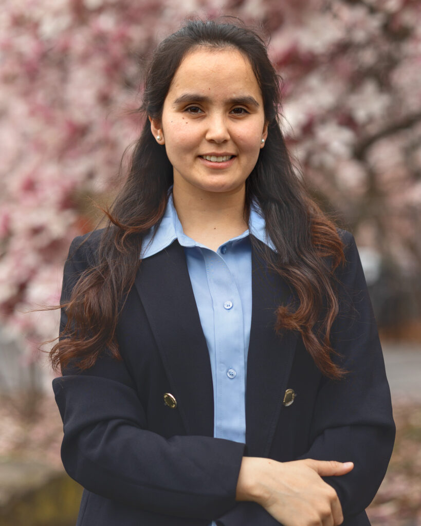 Tamana Ghaznawi smiles in front of a light pink flowering tree in the Arts Quad while wearing a black blazer and light blue button-up shirt.