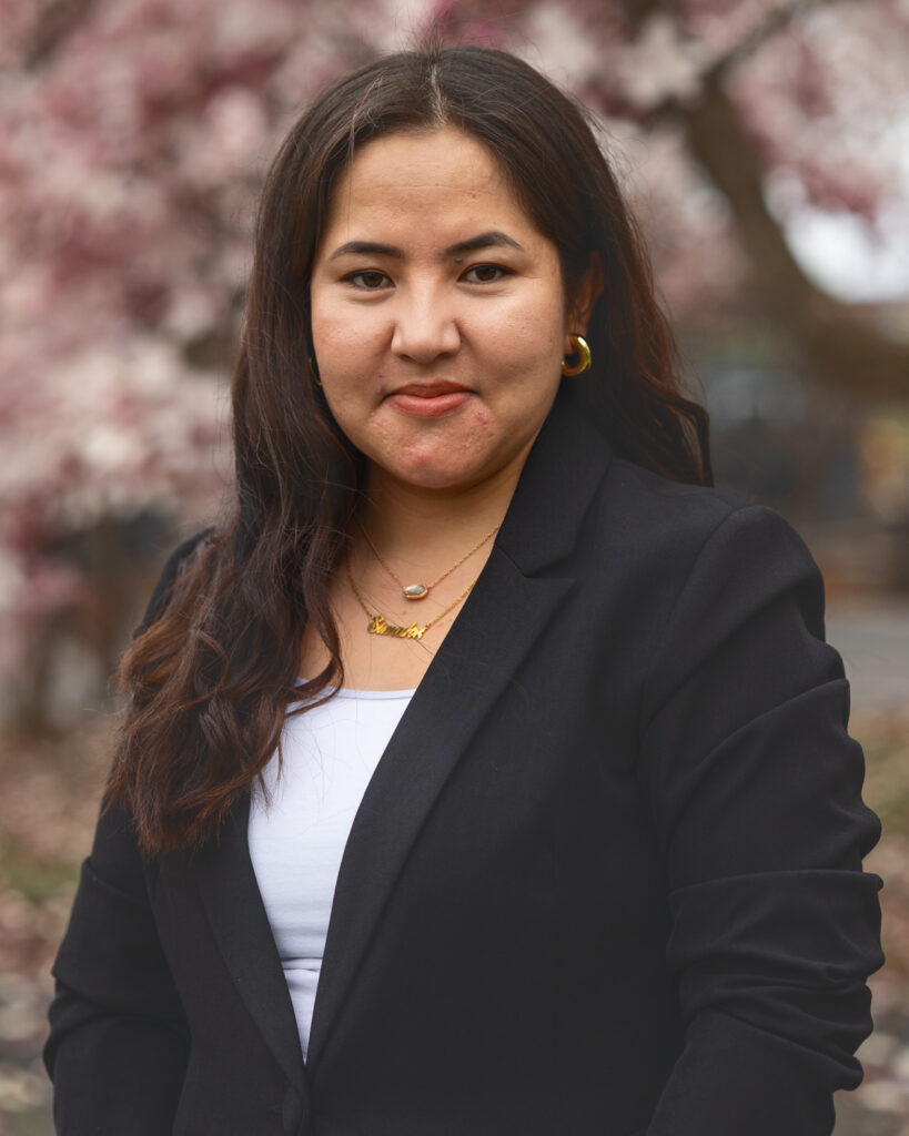 Simah Gul Sahnosh smiles in front of a light pink flowering tree in the Arts Quad while wearing a black blazer and white shirt.