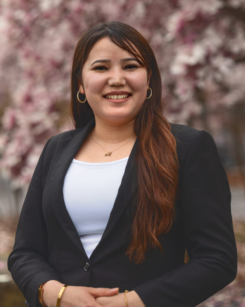 Khurshid Hussainy smiles in front of a light pink flowering tree in the Arts Quad while wearing a black blazer and white shirt.