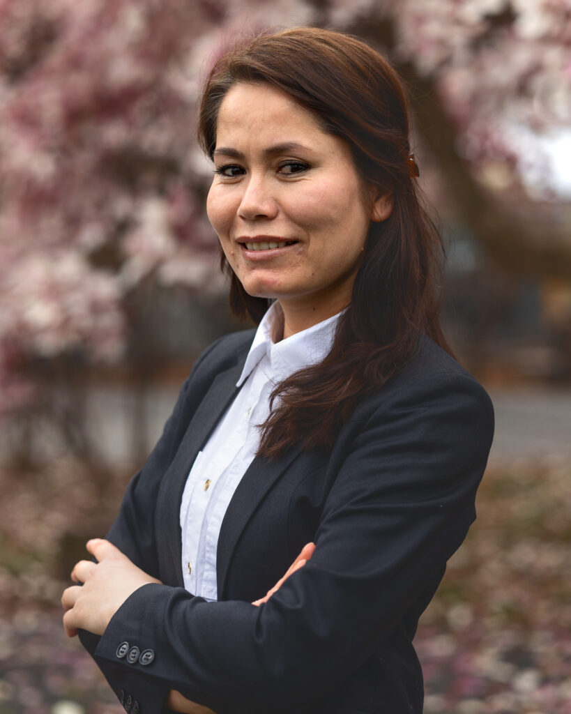 Khadija Monis smiles in front of a light pink flowering tree in the Arts Quad while wearing a black blazer and white shirt.