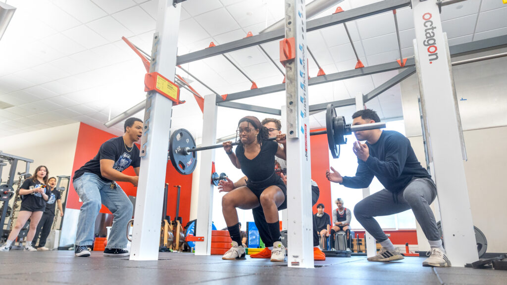 A member of the club squats weight while members around her spot her and cheer her on.