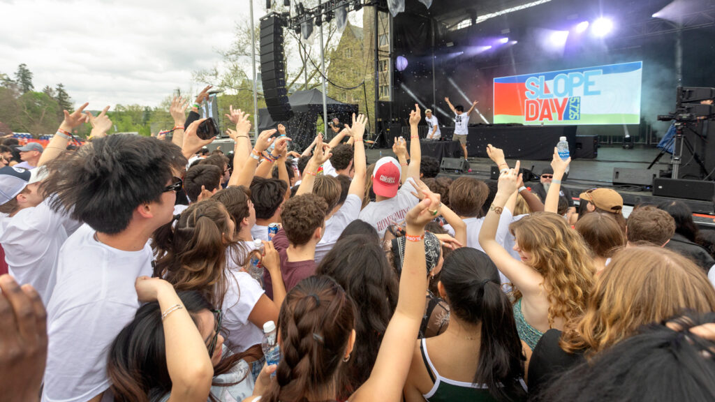 Students dance and hold their hands in the air while facing the stage. A screen that says "Slope Day" faces the crowd."