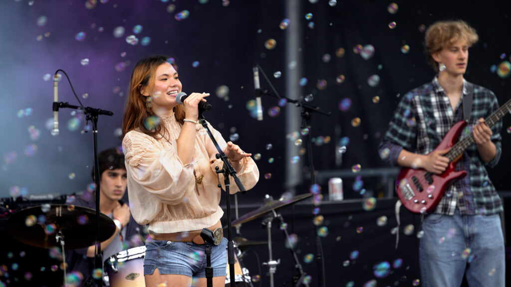 A female singer smiles while holding a microphone on stage standing next to an electric guitar player while bubbles float around them.