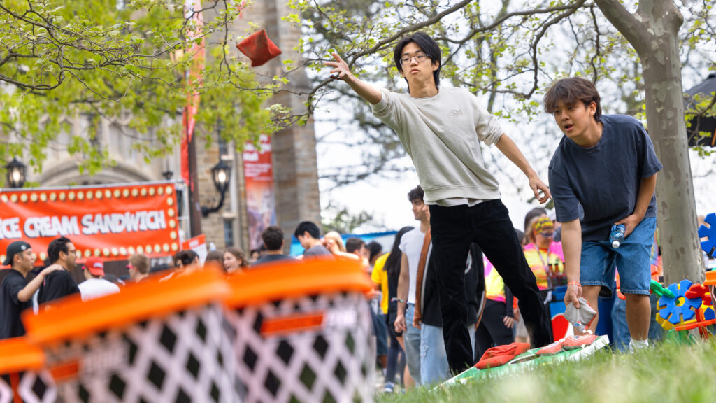 Two students stand next to each other playing cornhole. One tosses a red bag through the air.