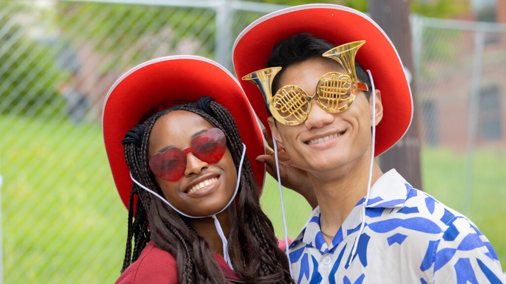 Two students in red cowboy hats smile for a photo. The student on the right is wearing glasses shaped like French horns.
