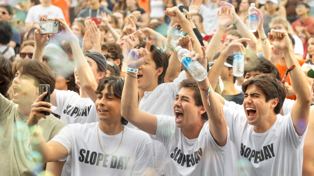 Students in the crowd yell with their hands in the air while wearing white t-shirts with "SLOPE DAY" on the front of them.