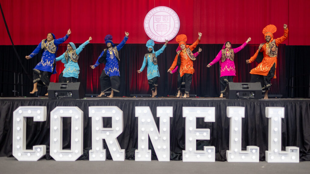 Seniors perform a cultural dance on stage during 2025 Senior Convocation in Barton Hall. In front of the stage "Cornell" is spelled out in large, white marquee light up letters.