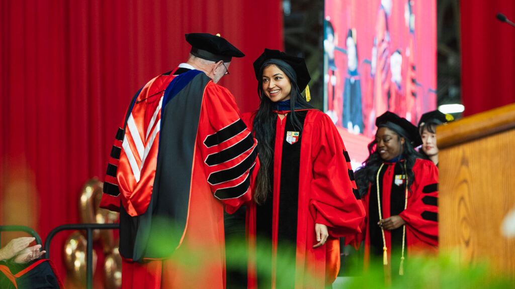 President Mike Kotlikoff, wearing his academic regalia, shakes hands with a smiling Ph.D. graduate, wearing her academic regalia, during the 2025 Ph.D. Recognition Ceremony.