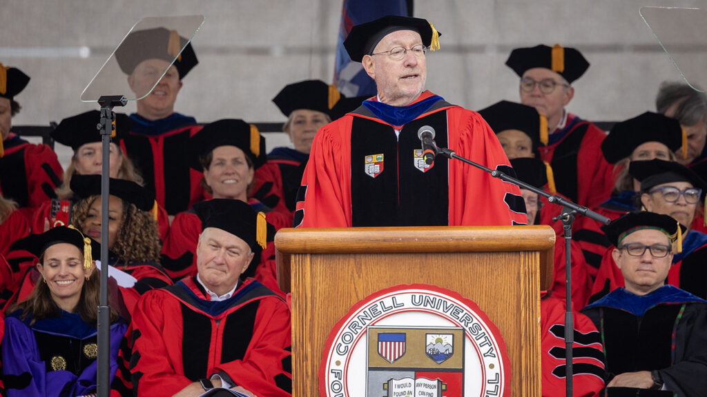 President Mike Kotlikoff, wearing academic regalia, speaks during commencement at Schoellkopf Field while faculty, also in academic regalia, sits behind him.