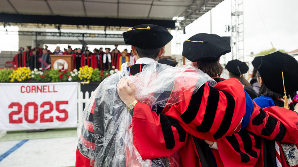Class of 2025 Ph.D. graduates wearing their academic regalia, and one also wearing a rain poncho, put their arms around each other during during commencement at Schoellkopf Field.