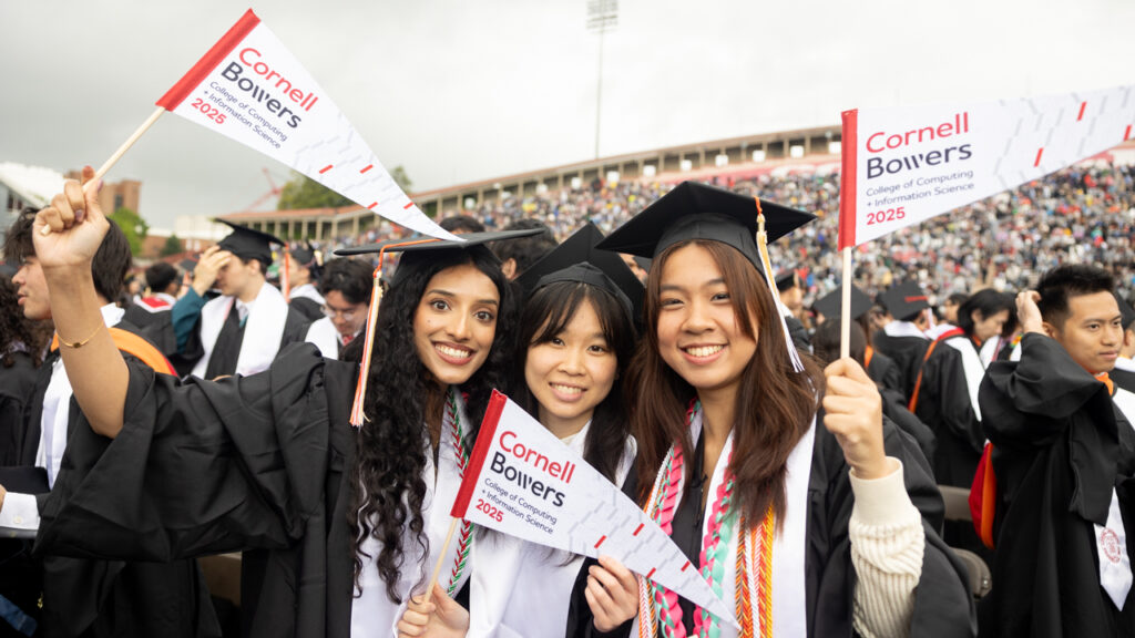 Three women graduates from the class of 2025 in their academic regalia smile and hold white and red flags representing the College of Computing and Information Science during commencement at Schoellkopf Field.