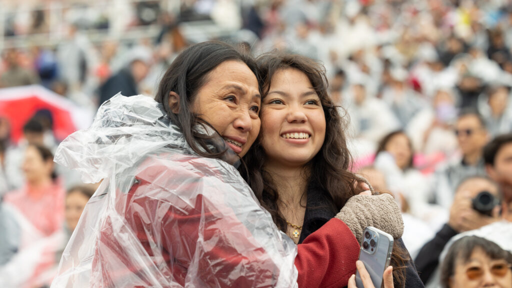 Two commencement attendees hug each other and smile at Schoellkopf Field.