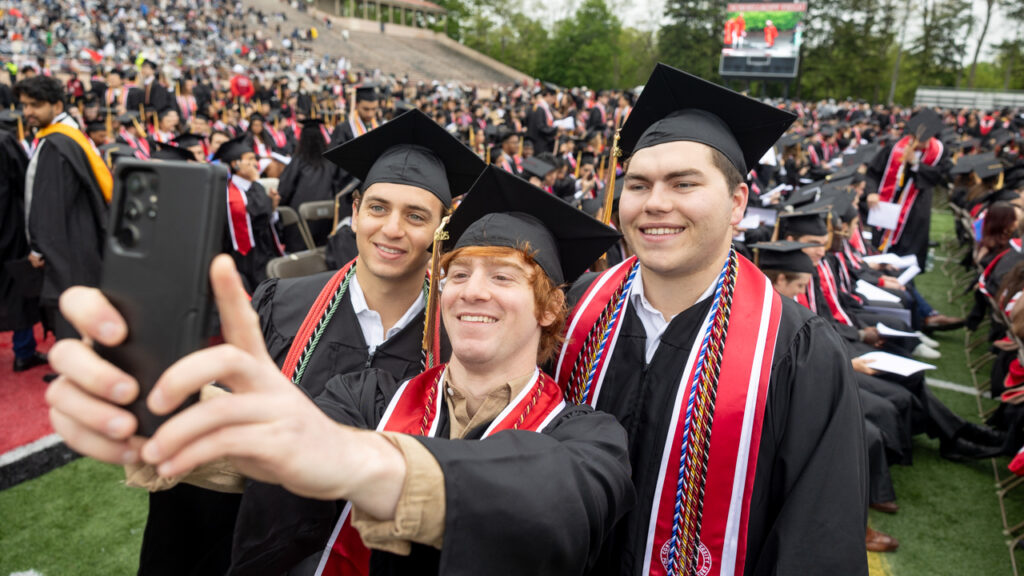 Three graduating men in academic regalia smile for a selfie during commencement at Schoellkopf Field.