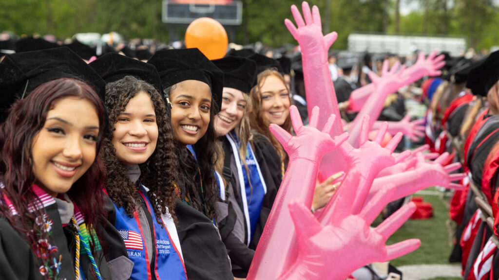 Graduates from the class of 2025 wear their academic regalia and stand in line smiling for the photo while holding pink inflatable hands to celebrate with.