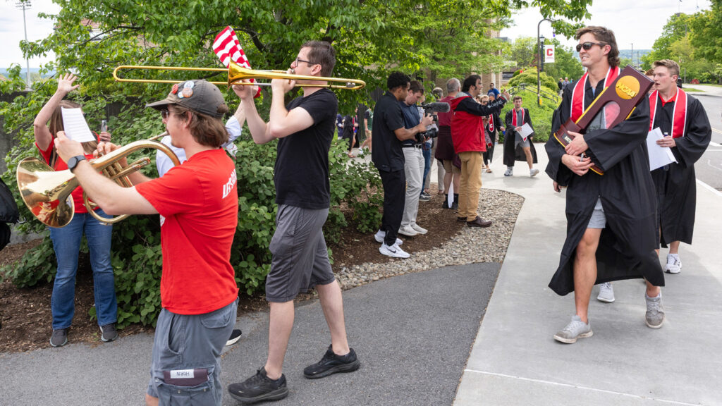 Two graduates, one holding the NCAA lacrosse trophy, follow students playing horn instruments in a parade