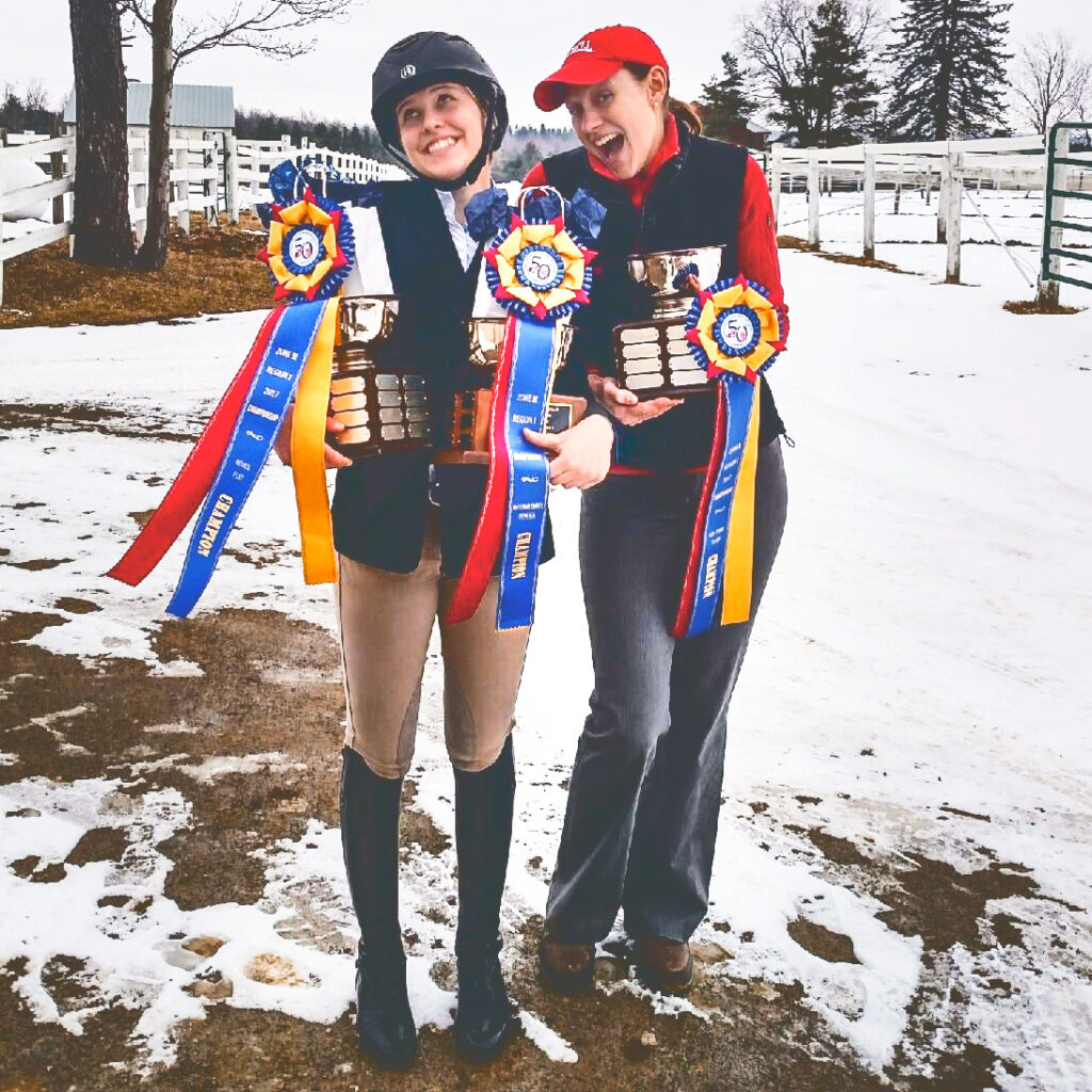 Joanna Novakovic and rider Claira Seely stand together outside after a show smiling and holding large trophies and champion ribbons.